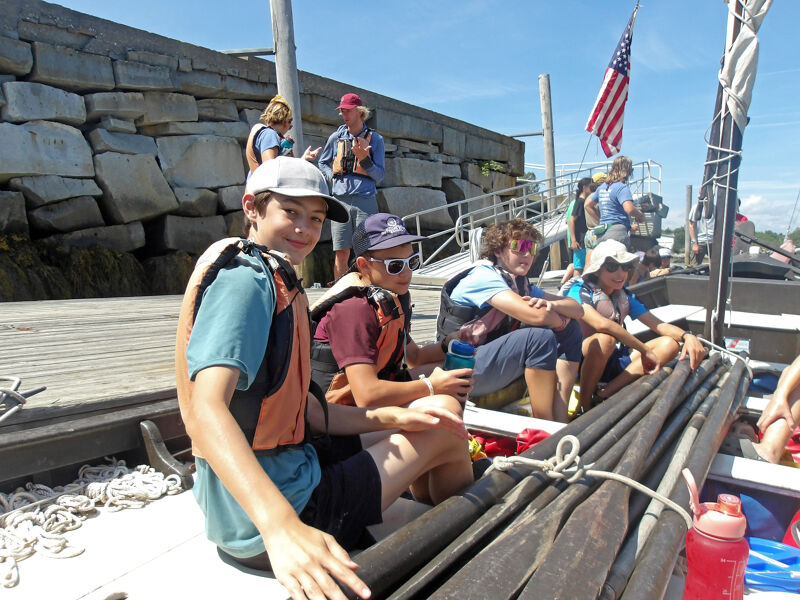 The image shows a group of young people on a boat, possibly a sailboat, with oars visible. They are wearing life jackets and casual clothing, suggesting a recreational activity. A stone wall is in the background, and an American flag is flying from the boat's mast. The overall scene indicates a sunny day and an outdoor adventure.